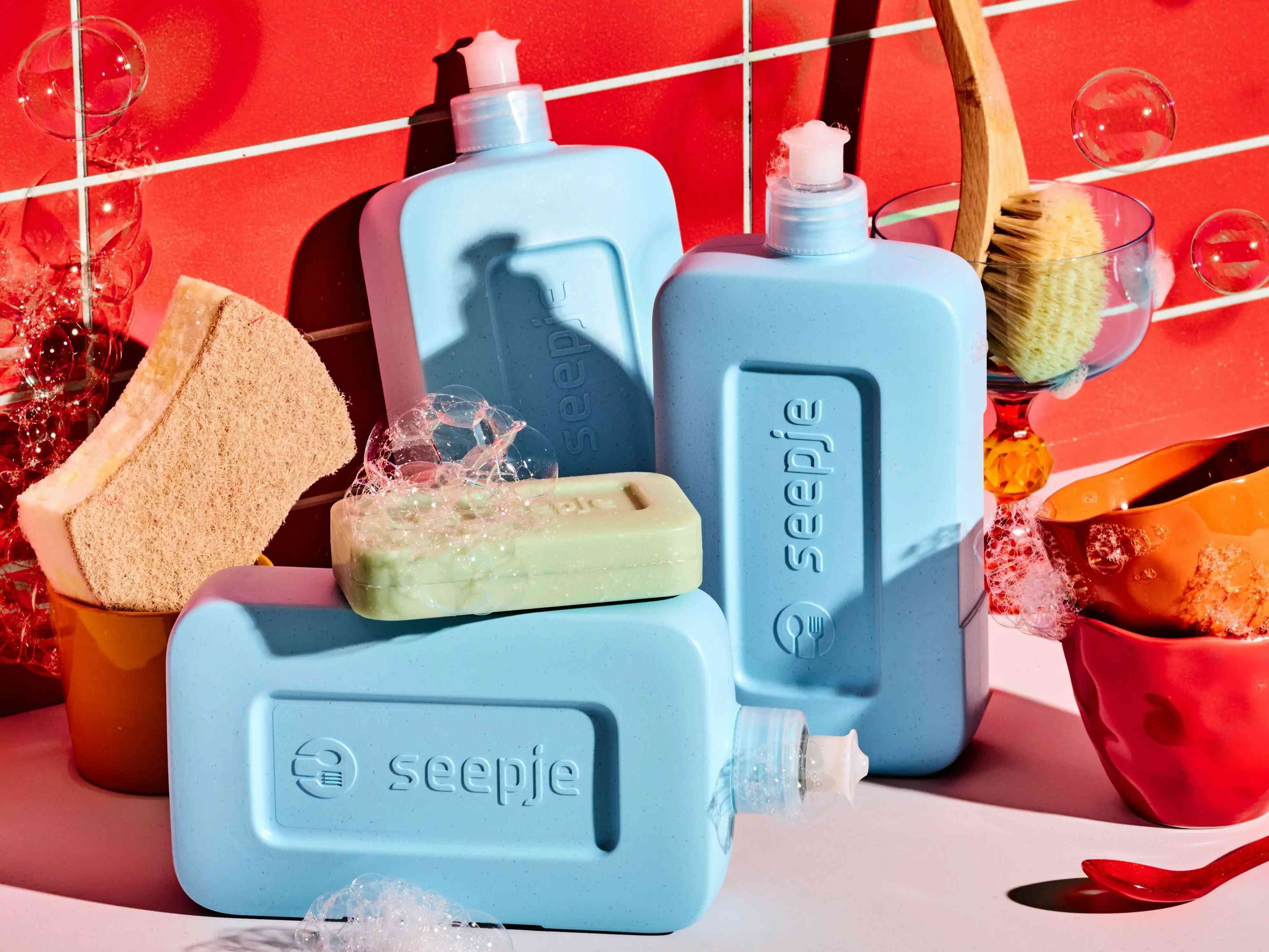 Soaply Dishwashing set with light blue refill bottles, a solid dish soap bar, sponge and brush, with bubbles against a red tiled background.